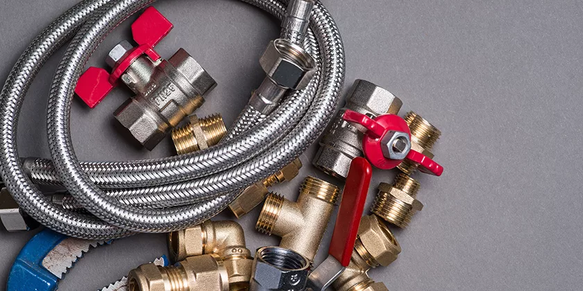 Assorted plumbing fittings, including brass connectors, valves with red handles, a flexible metal hose, and a blue wrench—essentials for espresso equipment maintenance—are arranged on a gray surface. © 2025 1st-line.com