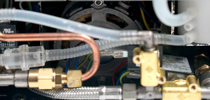 Close-up of internal components inside the Rocket R Cinquantotto (R58) Dual Boiler Espresso Machine, showing copper and plastic pipes, brass fittings, wiring, and part of a pump—illustrating its precise engineering. © 2025 1st-line.com