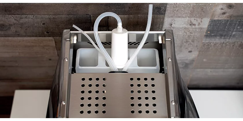 Top view of an ice maker machine with a metal grate, two white plastic ice trays, and clear tubes running from a central white cylinder—ideal for espresso equipment maintenance. The background features a textured, tiled wall. © 2025 1st-line.com
