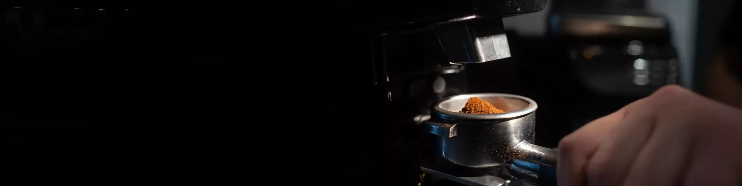 A close-up of a hand holding a portafilter filled with grounds from an espresso coffee grinder under an espresso machine, ready for brewing. The dark background highlights the coffee preparation process. © 2025 1st-line.com
