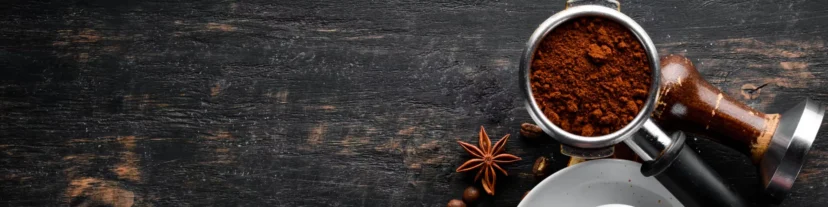 A portafilter filled with ground coffee sits on a dark wooden surface near a tamper, anise stars, coffee beans, and part of a white cup—essential parts for anyone passionate about espresso machines. © 2025 1st-line.com