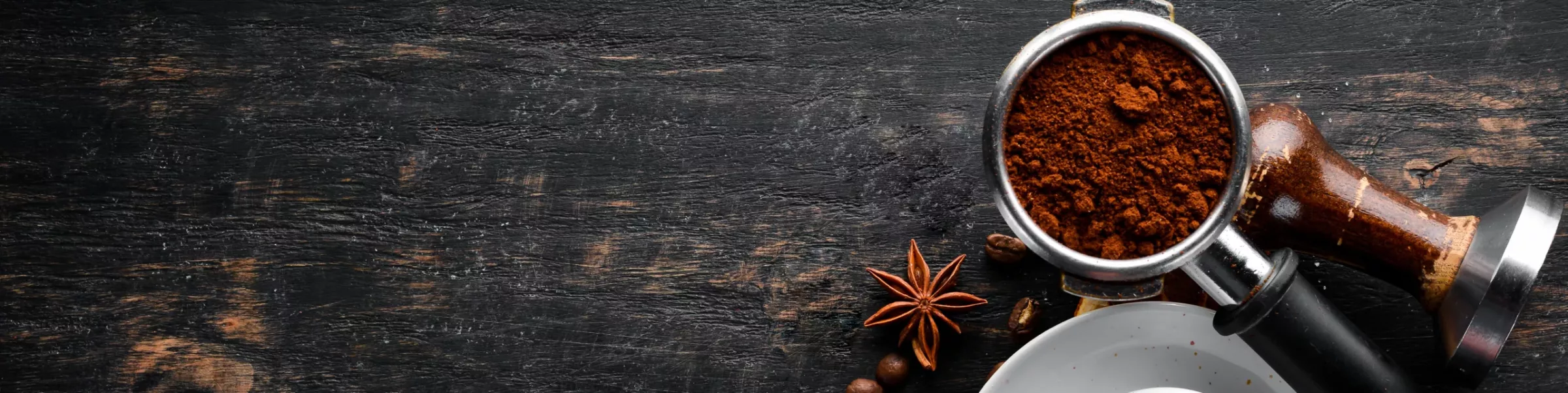 A portafilter filled with ground coffee sits on a dark wooden surface near a tamper, anise stars, coffee beans, and part of a white cup—essential parts for anyone passionate about espresso machines. © 2025 1st-line.com
