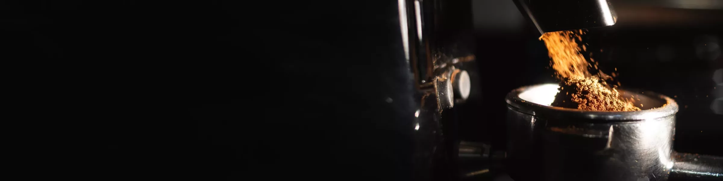 Ground coffee is being dispensed from a semi-automatic grinder into a metal portafilter, with a dark background emphasizing the coffee grounds and equipment. © 2025 1st-line.com