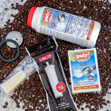 The Maintenance Care Package, including Puly Caff, a syringe kit, brush, filter, and detergent sachets, is neatly arranged on marble among coffee beans—ideal for easy espresso machine upkeep. © 2025 1st-line.com