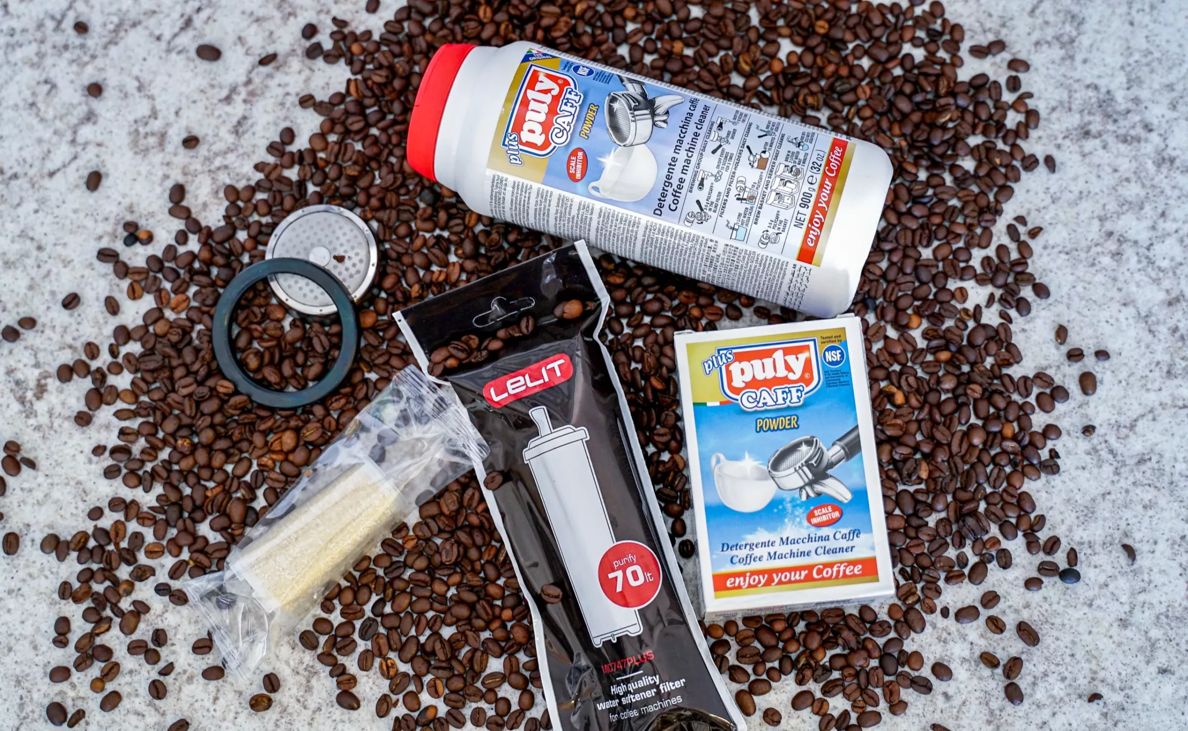 The Maintenance Care Package, including Puly Caff, a syringe kit, brush, filter, and detergent sachets, is neatly arranged on marble among coffee beans—ideal for easy espresso machine upkeep. © 2025 1st-line.com
