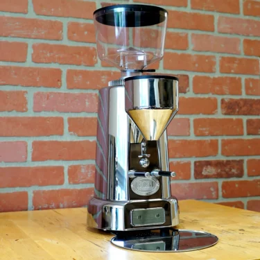 A used SIDEWALK SALE - ECM V-Titan 64 Espresso Grinder (USED E209) with a clear hopper and round drip tray sits on a wooden table before a red brick wall, showcasing ECM’s signature style and precision in stainless steel. © 2025 1st-line.com