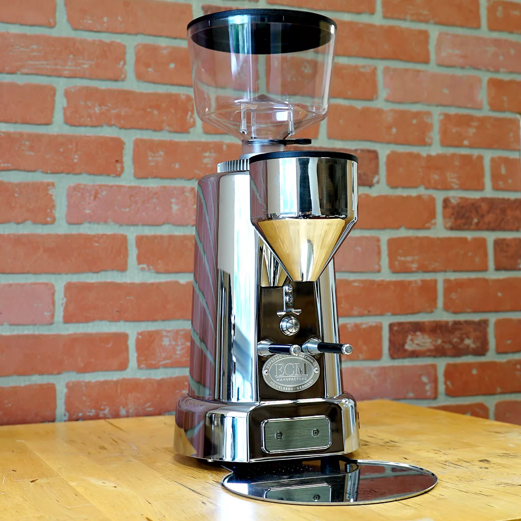 A used SIDEWALK SALE - ECM V-Titan 64 Espresso Grinder (USED E209) with a clear hopper and round drip tray sits on a wooden table before a red brick wall, showcasing ECM’s signature style and precision in stainless steel. © 2025 1st-line.com