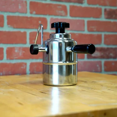 The SIDEWALK SALE - Stovetop Steamer, a shiny stainless steel coffee maker with black handles, sits on a wooden table against a red brick wall with white mortar. © 2025 1st-line.com