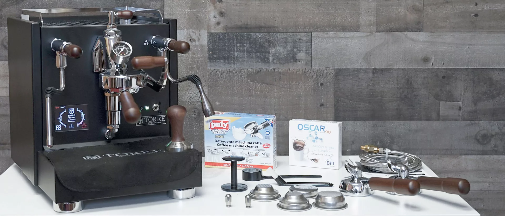 A black Torre Peppina EVO Premium V1 Dual Boiler Espresso Machine w/ FC with wooden handles sits on a white surface beside portafilters, filter baskets, a tamper, cleaning items, and metal hoses; a wooden wall forms the background. © 2025 1st-line.com