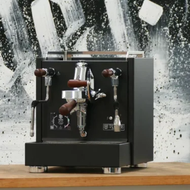 A Torre Peppina EVO Premium V1 Dual Boiler Espresso Machine w/ FC in sleek black with chrome and wood accents sits on a wooden counter, set against an abstract black-and-white painted wall. © 2025 1st-line.com