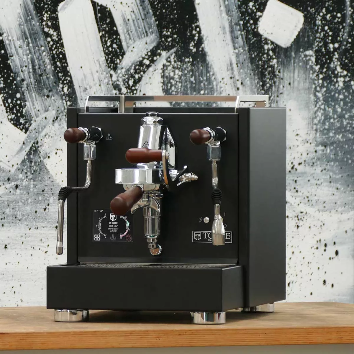 A Torre Peppina EVO Premium V1 Dual Boiler Espresso Machine w/ FC in sleek black with chrome and wood accents sits on a wooden counter, set against an abstract black-and-white painted wall. © 2025 1st-line.com
