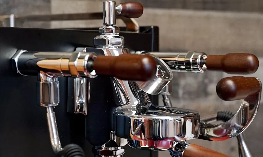 Close-up of the Torre Peppina EVO Premium V1 Dual Boiler Espresso Machine w/ FC, featuring a shiny chrome finish, wooden handles, and a pressure gauge, set against a blurred background. © 2025 1st-line.com