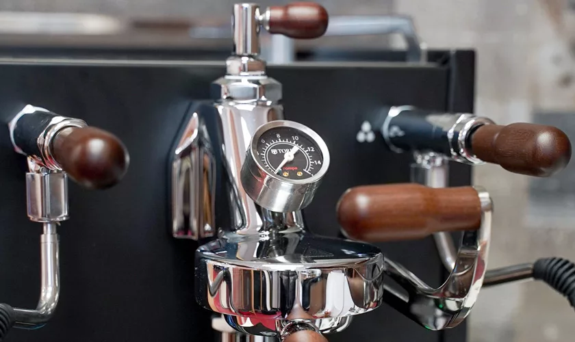 Close-up of the Torre Peppina EVO Premium V1 Dual Boiler Espresso Machine w/ FC, featuring wood-accented handles and a central pressure gauge. The machines sleek design stands out against a softly blurred background. © 2025 1st-line.com