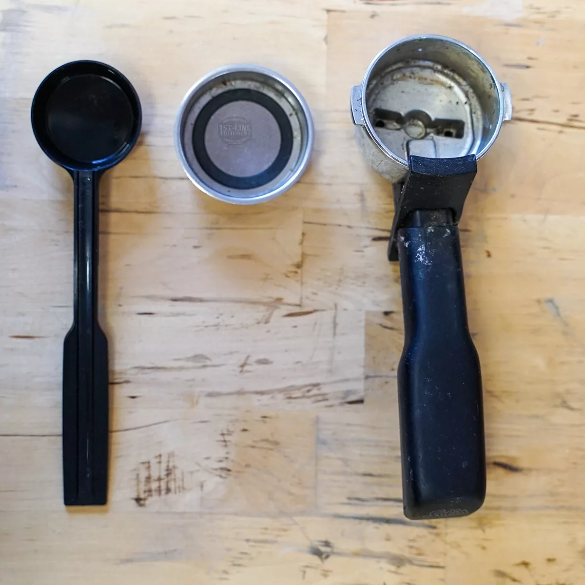 A black plastic scoop, metal filter basket, and a Solis Crema SL 90 (FOR PARTS ONLY) (E109) portafilter with black handle are arranged on wood—great picks for your SIDEWALK SALE collection. © 2025 1st-line.com