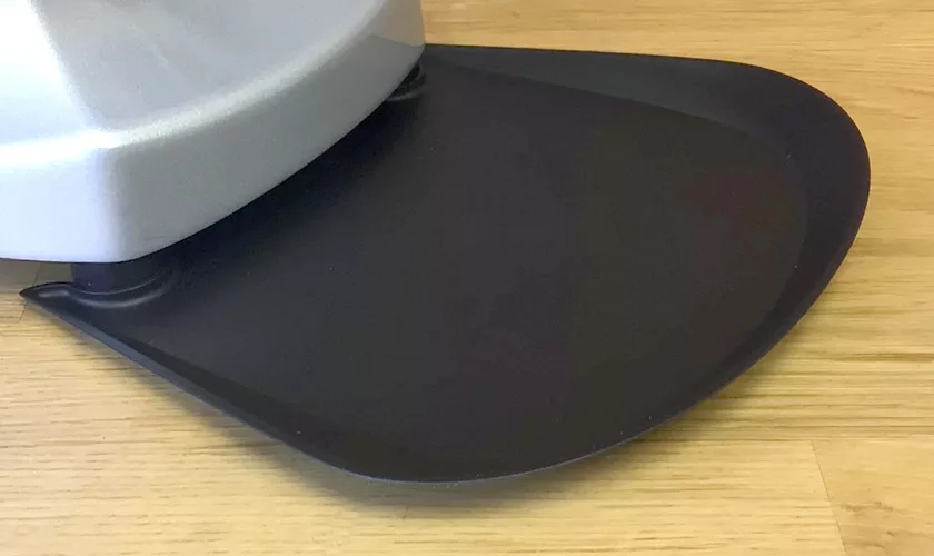 A close-up of a black plastic drip tray attached to the base of a silver Mazzer Super Jolly V UP Espresso Coffee Grinder, placed on a light wooden surface. © 2025 1st-line.com