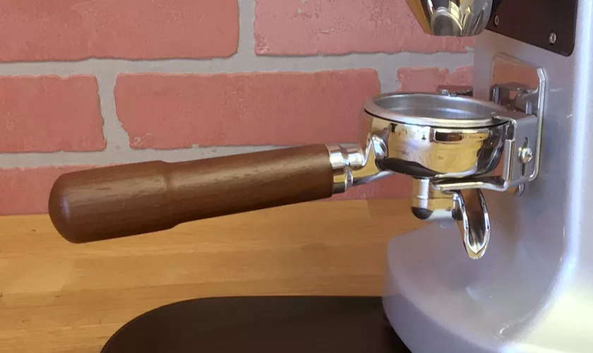 A close-up of a coffee portafilter with a wooden handle attached to an espresso machine on a wooden countertop, with the Mazzer Super Jolly V UP Espresso Coffee Grinder visible in the background against a brick wall. © 2025 1st-line.com