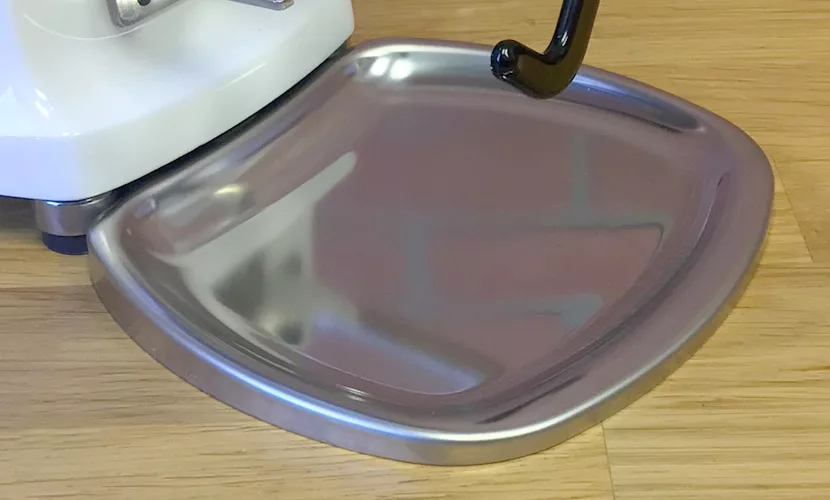 A close-up of a shiny, rectangular stainless steel tray attached to a white Mazzer Mini B Electronic Time-Based Espresso Coffee Grinder, placed on a light wooden surface. © 2025 1st-line.com