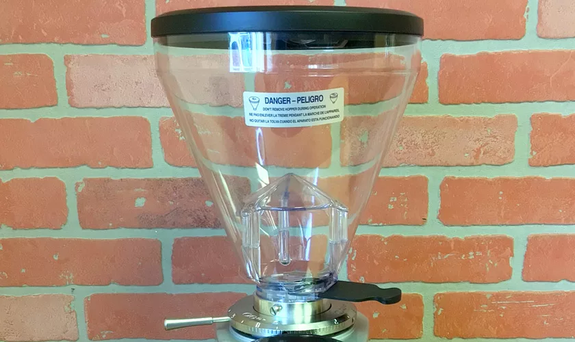 A close-up of the Mazzer Super Jolly V UP Espresso Coffee Grinder shows its large clear hopper with a black lid and a visible warning label, set against a red brick wall backdrop. © 2025 1st-line.com