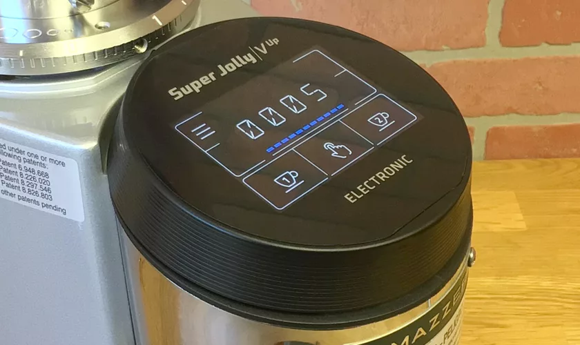 A close-up of the Mazzer Super Jolly V UP Espresso Coffee Grinder’s digital control panel, featuring touch-sensitive buttons and a digital display, set against a wooden surface and brick wall background. © 2025 1st-line.com