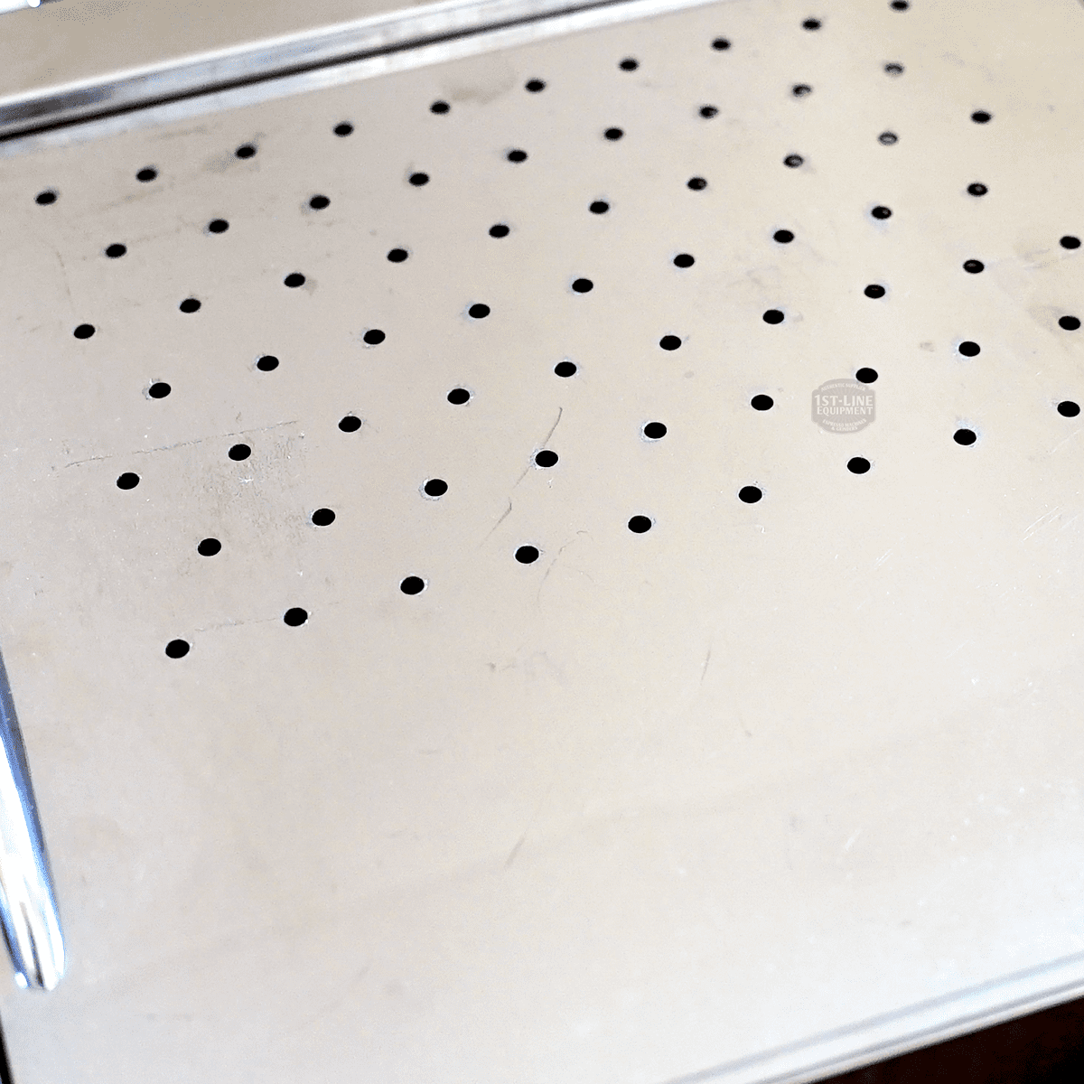 A close-up of the metal drip tray, featuring evenly spaced round holes, from the SIDEWALK SALE - VBM Domobar Super HX Switchable Espresso Machine OPEN BOX (E110), showing some scratches and reflections. © 2025 1st-line.com