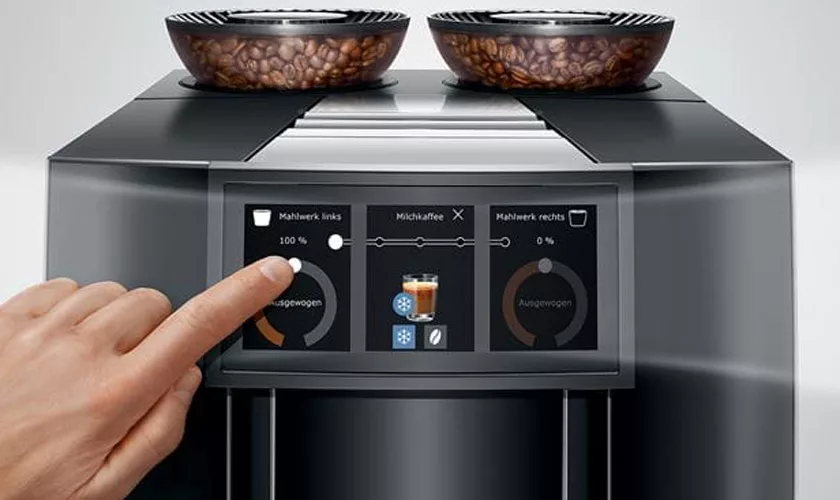 A hand touches the touchscreen of the Jura GIGA 10 Super-Automatic Espresso Machine, featuring two coffee bean compartments on top and a display showing coffee settings and options in German. © 2025 1st-line.com