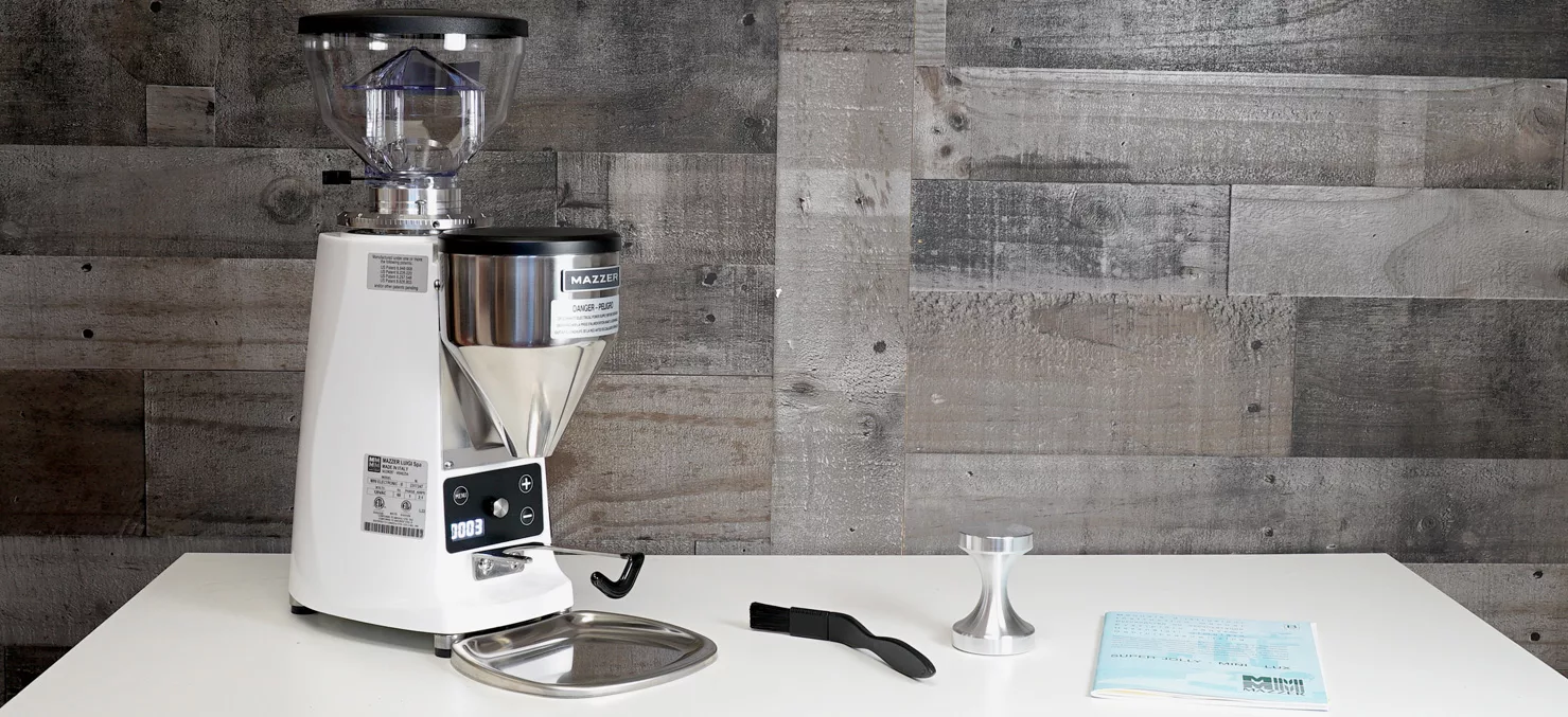 A Mazzer Mini B Electronic Time-Based Espresso Coffee Grinder sits on a white table with a tamper, brush, and instruction manual beside it against a gray wooden wall background. © 2025 1st-line.com