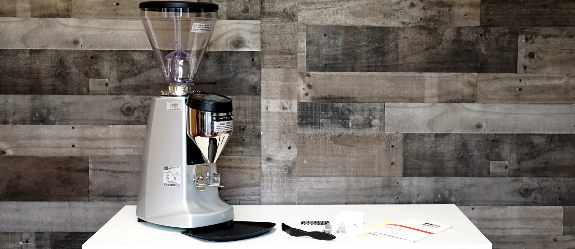 A Mazzer Super Jolly V UP Espresso Coffee Grinder is placed on a white table against a wooden plank wall, with accessories such as a brush, spoon, cleaning tool, and instruction manuals beside it. © 2025 1st-line.com