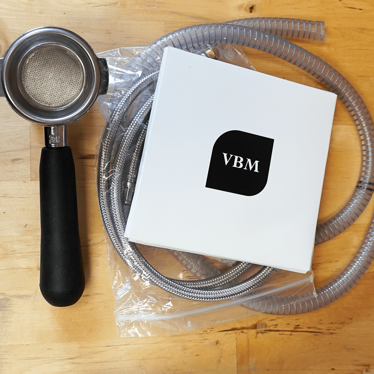 A portafilter with a black handle, a flexible braided hose, and a white VBM box are displayed on wood—ideal with the SIDEWALK SALE - VBM Domobar Super HX Switchable Espresso Machine OPEN BOX (E110). © 2025 1st-line.com