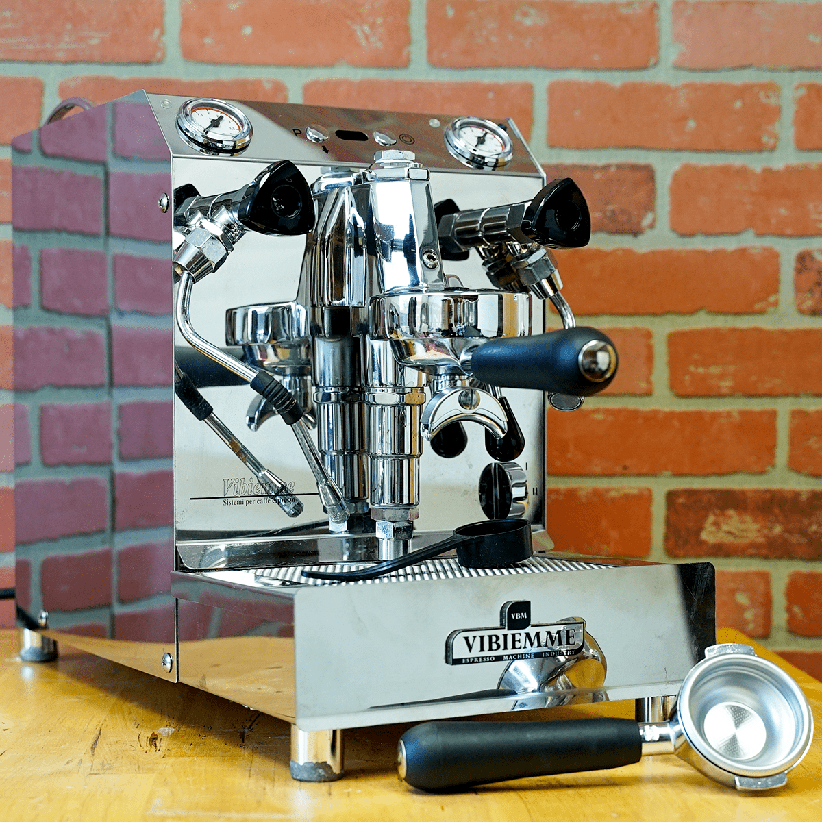 A shiny stainless steel SIDEWALK SALE – VBM Super Electronic Dual Boiler Espresso Machine - direct connect only (E114) sits on a wooden surface before a red brick wall, displaying multiple knobs, levers, and its portafilter in front. © 2025 1st-line.com