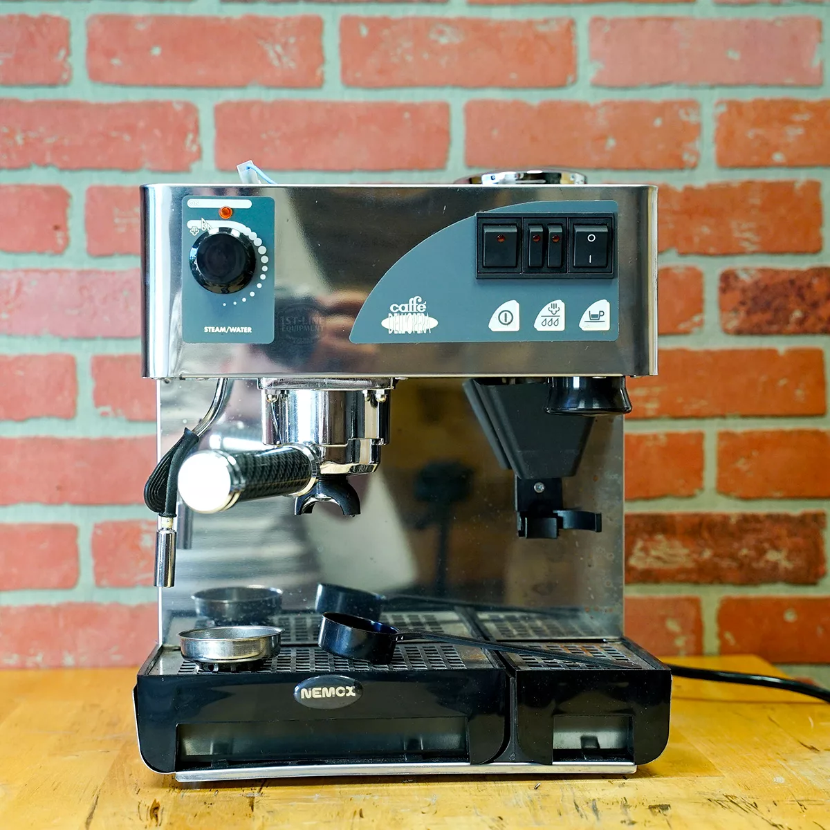 A SIDEWALK SALE Nemo Professional Caffe DellOpera (FOR PARTS ONLY) (E012) sits on a wooden countertop before a red brick wall, featuring stainless steel, black accents, buttons, steam wand, and portafilter—perfect for parts seekers. © 2025 1st-line.com