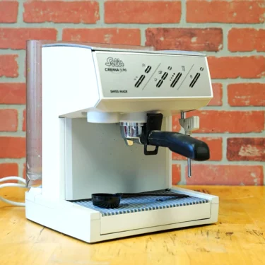 A SIDEWALK SALE Solis Crema SL 90 espresso machine (FOR PARTS ONLY, E109) sits on a wooden surface by a red brick wall, featuring black handles and a small black scoop on the drip tray—great for parts or your next find!. © 2025 1st-line.com