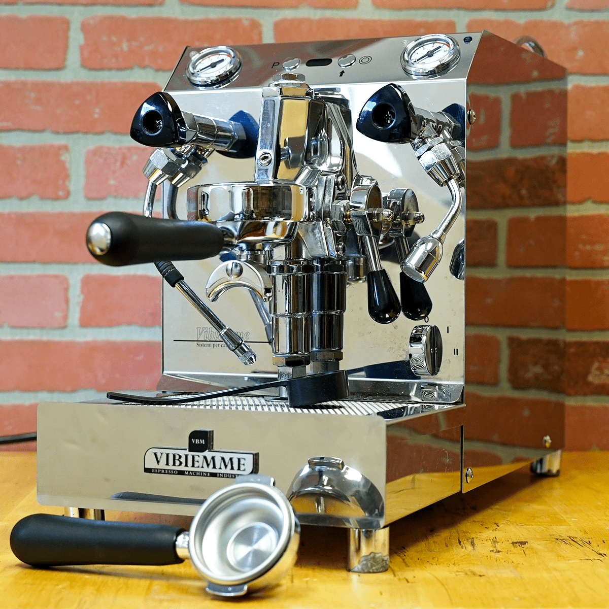 A SIDEWALK SALE – VBM Super Electronic Dual Boiler Espresso Machine (direct connect, E114) sits on a wooden counter before a brick wall, its detachable portafilter on the drip tray—ideal for coffee lovers. © 2025 1st-line.com