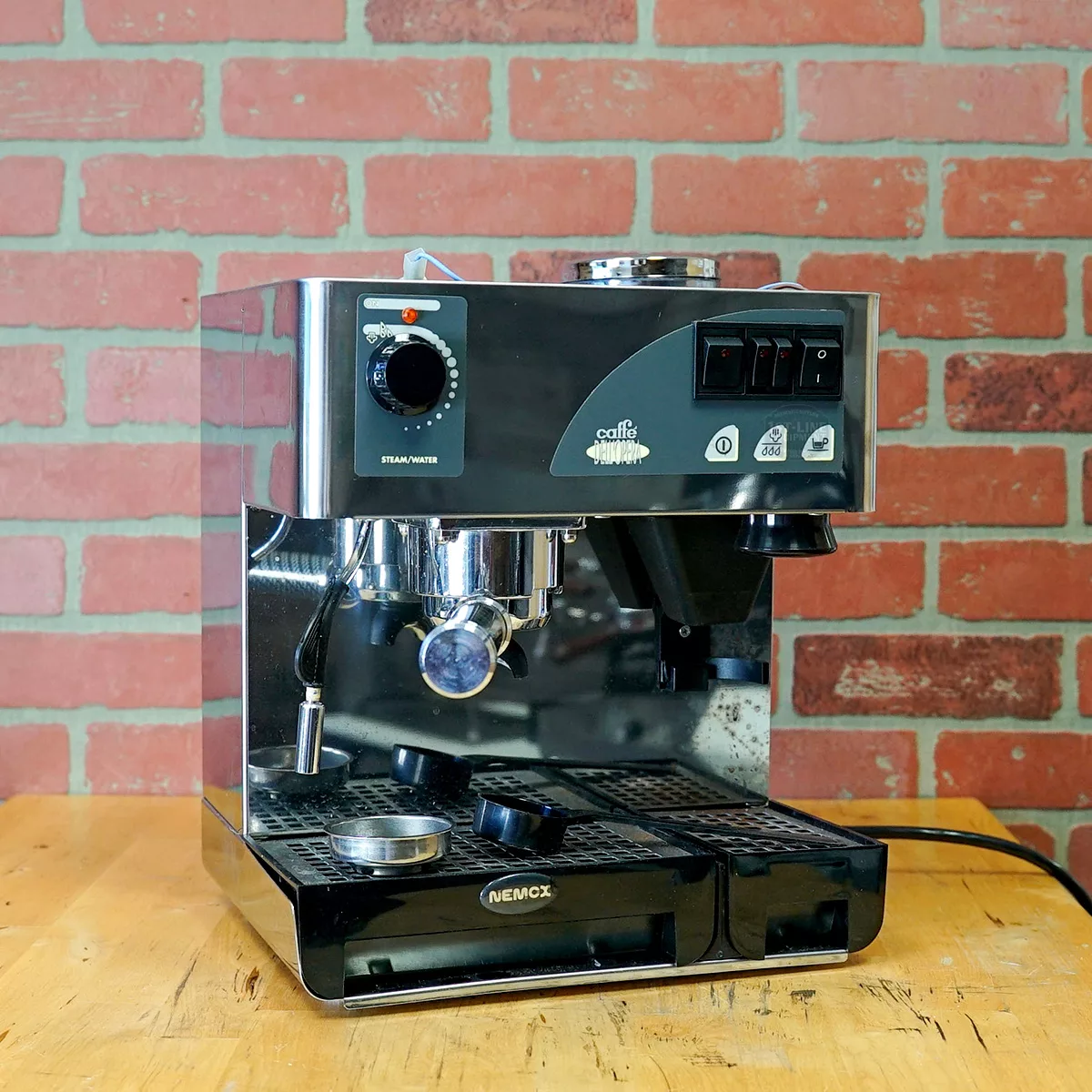 A stainless steel Nemox espresso machine sits on a wooden surface in front of a red brick wall, featuring buttons, a portafilter, and a steam wand. © 2025 1st-line.com
