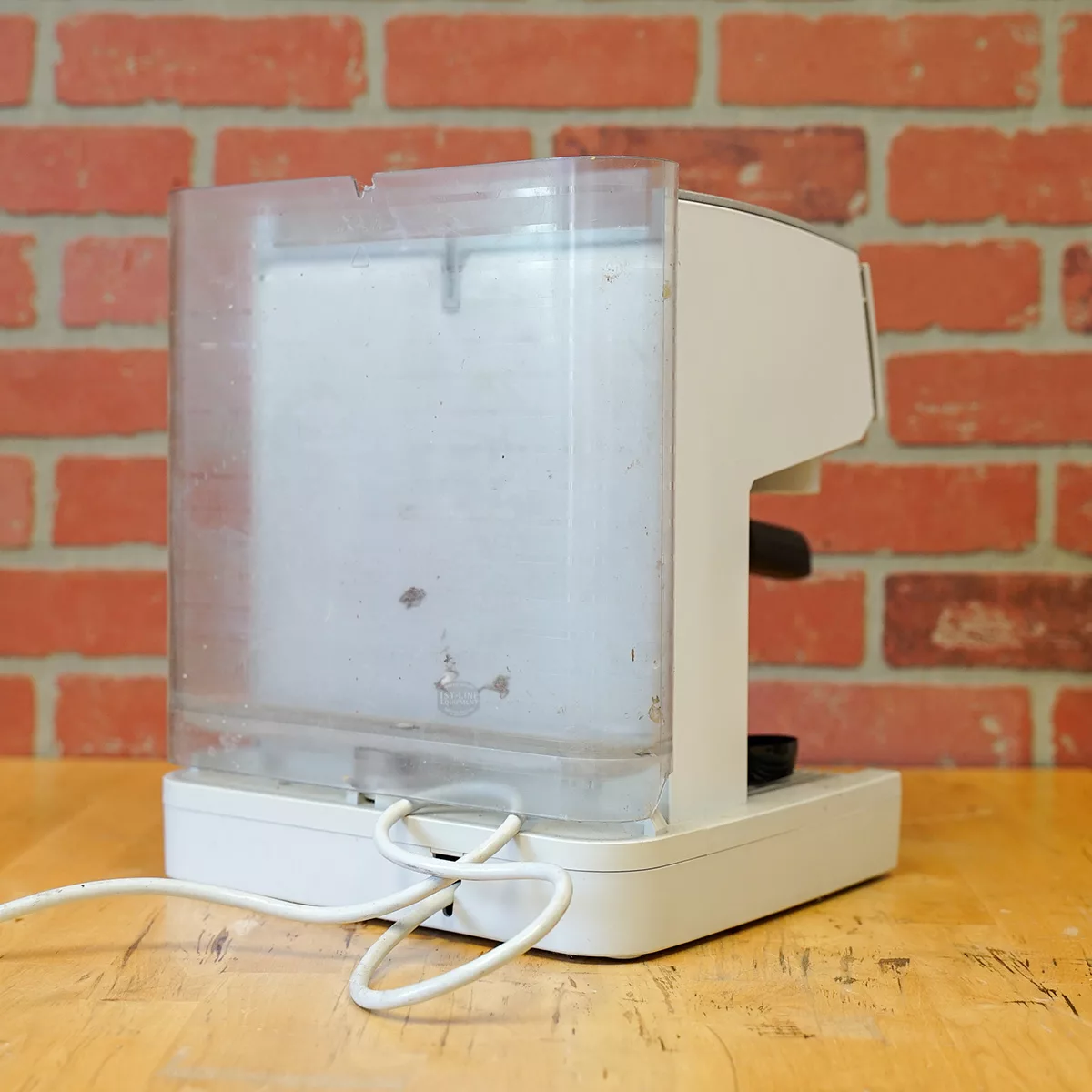 A white Solis Crema SL 90 espresso machine (FOR PARTS ONLY) with a translucent water tank sits unplugged, cord coiled, on a wooden table by a red brick wall—ready for the SIDEWALK SALE. Model: E109. © 2025 1st-line.com