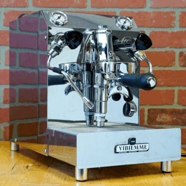 An open box SIDEWALK SALE - VBM Domobar Super HX Switchable Espresso Machine (E110) rests on a wooden counter with dual spouts and black handles, set against a red brick wall background. © 2025 1st-line.com