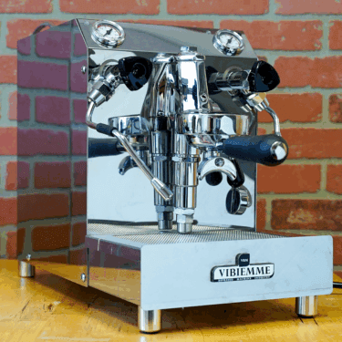An open box SIDEWALK SALE - VBM Domobar Super HX Switchable Espresso Machine (E110) rests on a wooden counter with dual spouts and black handles, set against a red brick wall background. © 2025 1st-line.com