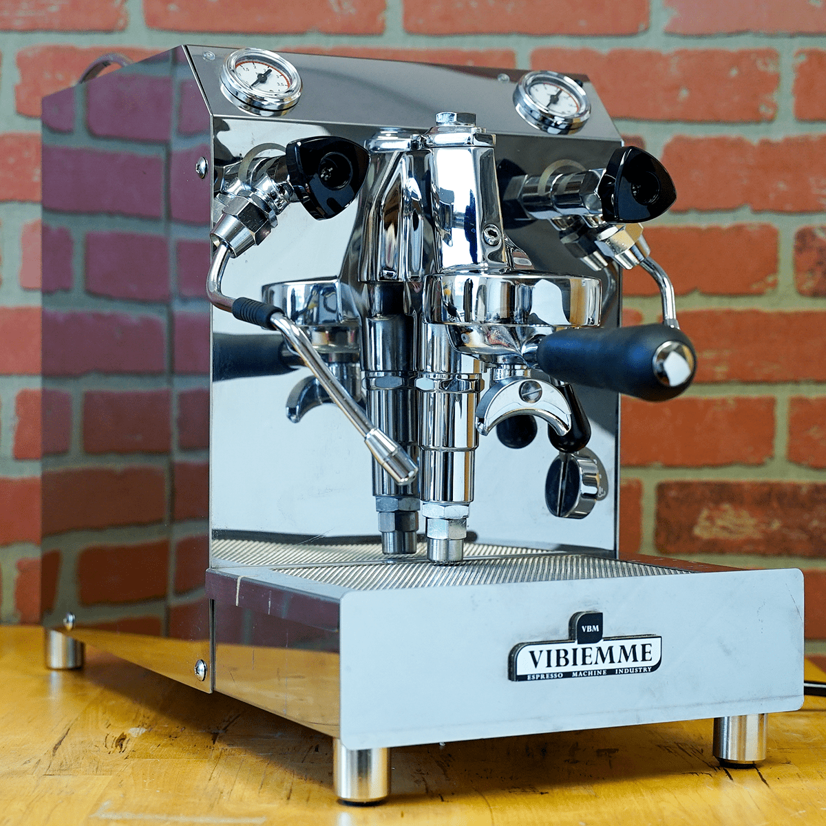 An open box SIDEWALK SALE - VBM Domobar Super HX Switchable Espresso Machine (E110) rests on a wooden counter with dual spouts and black handles, set against a red brick wall background. © 2025 1st-line.com