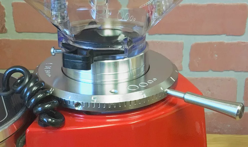 Close-up of a Mazzer Mini A Electronic Time-Based Espresso Coffee Grinder with a red base, featuring the numbered grind size adjustment ring, metal lever, and part of the coiled black cable. Brick wall in the background. © 2025 1st-line.com