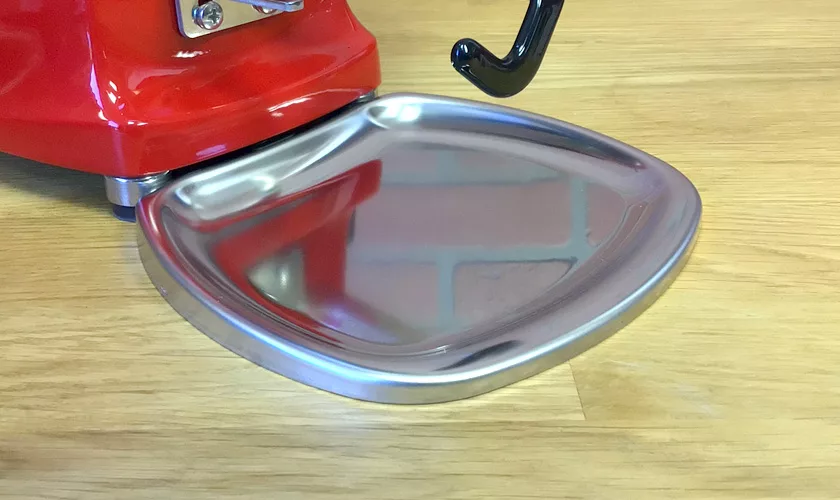 Close-up of a shiny, empty metal tray attached to the base of the red Mazzer Mini A Electronic Time-Based Espresso Coffee Grinder on a wooden surface. © 2025 1st-line.com