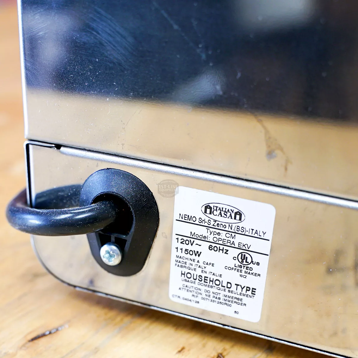 Close-up of a SIDEWALK SALE Nemo Professional Caffe DellOpera (FOR PARTS ONLY) (E012) metal appliance with power cord plugged in and specs label, displayed on a wooden surface. © 2025 1st-line.com