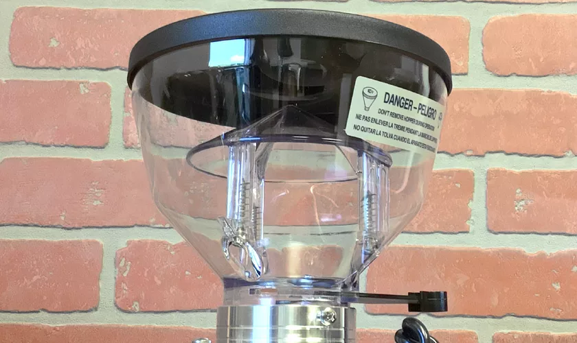 Close-up of a transparent hopper with black lid on a Mazzer Mini B Electronic Time-Based Espresso Coffee Grinder, attached to a metal body. A warning label is visible, and the background shows a red brick patterned wall. © 2025 1st-line.com