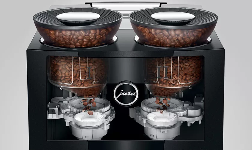 Close-up of the Jura GIGA 10 Super-Automatic Espresso Machine, showing two transparent bean containers on top with coffee beans dispensing into separate internal grinders. © 2025 1st-line.com
