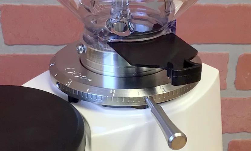 Close-up of the Mazzer Mini B Electronic Time-Based Espresso Coffee Grinder’s numbered adjustment dial and metal lever, set against a red and beige brick wall background. © 2025 1st-line.com