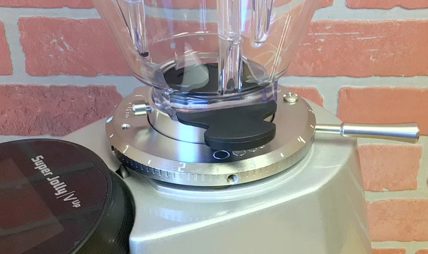 Close-up of the Mazzer Super Jolly V UP Espresso Coffee Grinder’s adjustment dial, featuring a clear plastic hopper, black handle, and metal lever; a brick wall appears in the background. © 2025 1st-line.com