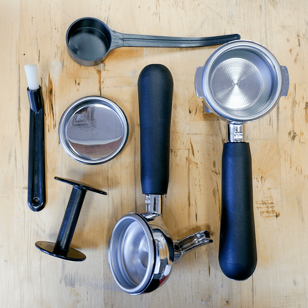Espresso machine accessories—two portafilters, coffee scoop, tamper, and more with sleek black handles—on wood surface, perfect for the SIDEWALK SALE VBM Super Electronic Dual Boiler Espresso Machine (direct connect only, E114). © 2025 1st-line.com
