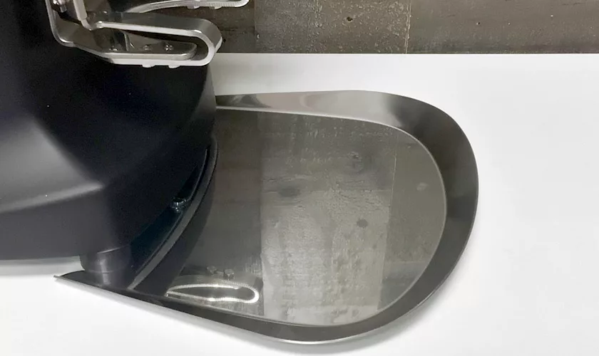 A close-up of a metal drip tray attached to the base of a Mazzer Super Jolly V Pro Espresso Coffee Grinder, shown on a white surface with a gray wall in the background. © 2025 1st-line.com