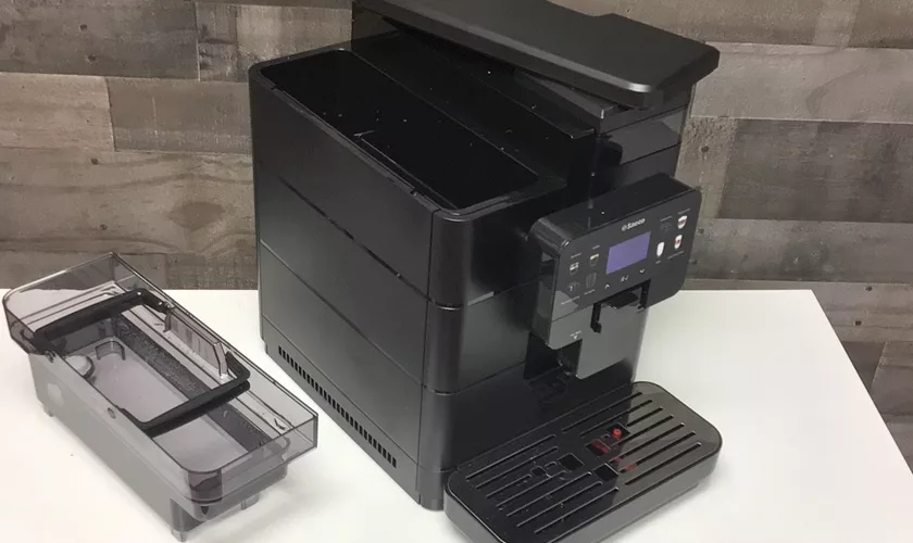 A DISCONTINUED Saeco New Royal OTC Super-Automatic Espresso Machine sits on a white surface beside its removed clear water reservoir, showcasing its control panel and drip tray against a wooden patterned wall. © 2025 1st-line.com