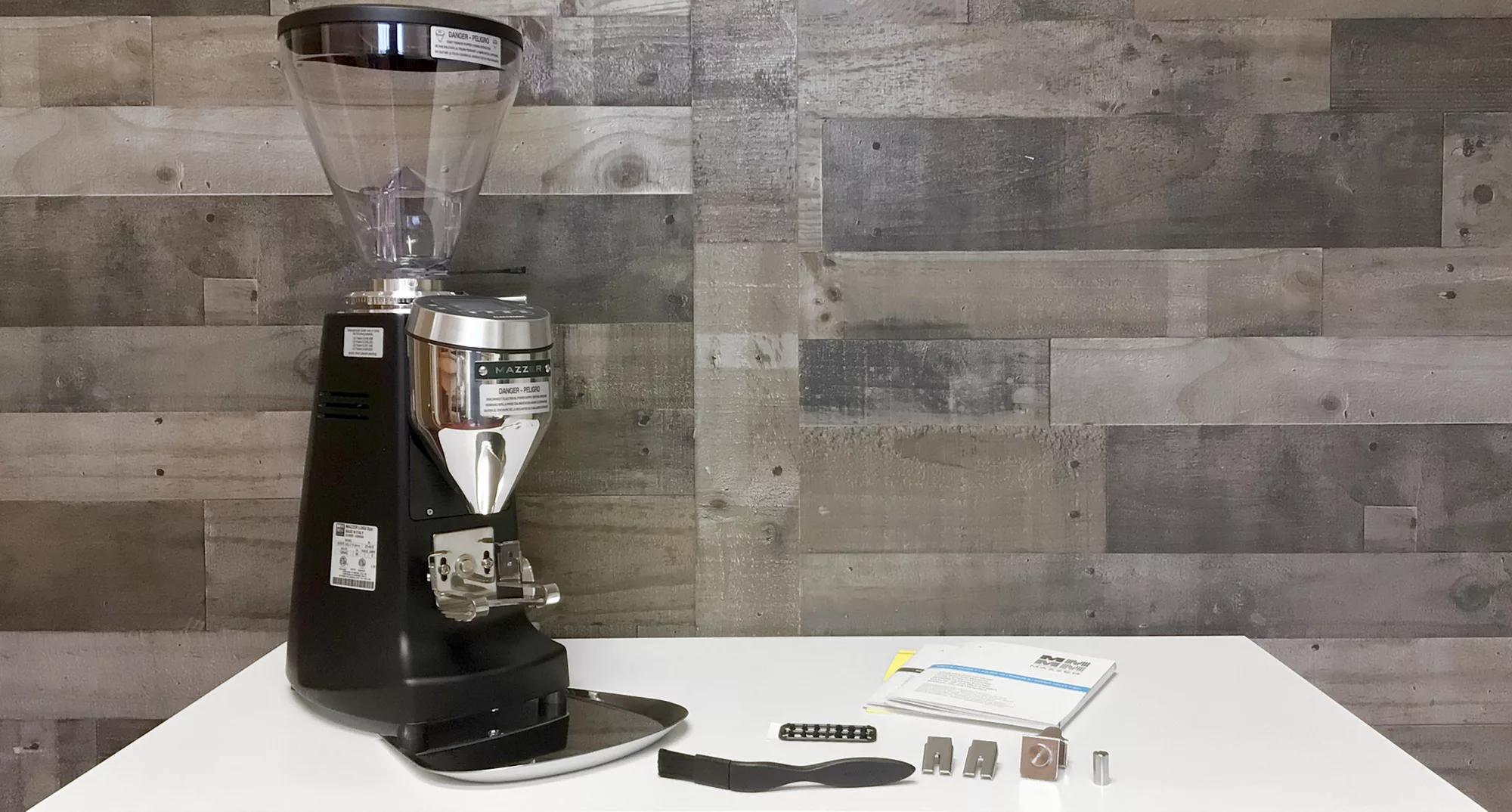 A Mazzer Super Jolly V Pro Espresso Coffee Grinder in black and silver sits on a white table with a black brush, metal parts, and a manual, set against a gray wooden wall background. © 2025 1st-line.com