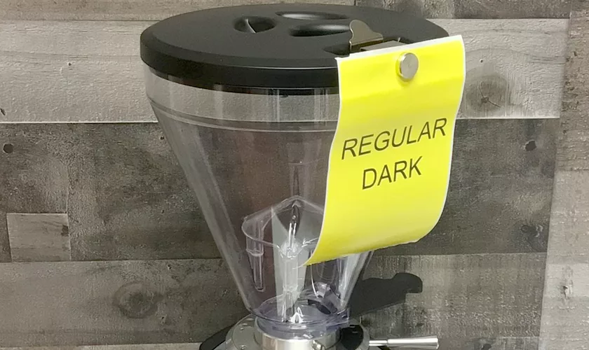 A Mazzer Super Jolly V Pro Espresso Coffee Grinder with a clear hopper, black lid, and a yellow REGULAR DARK sign is set against a wooden wall background. © 2025 1st-line.com