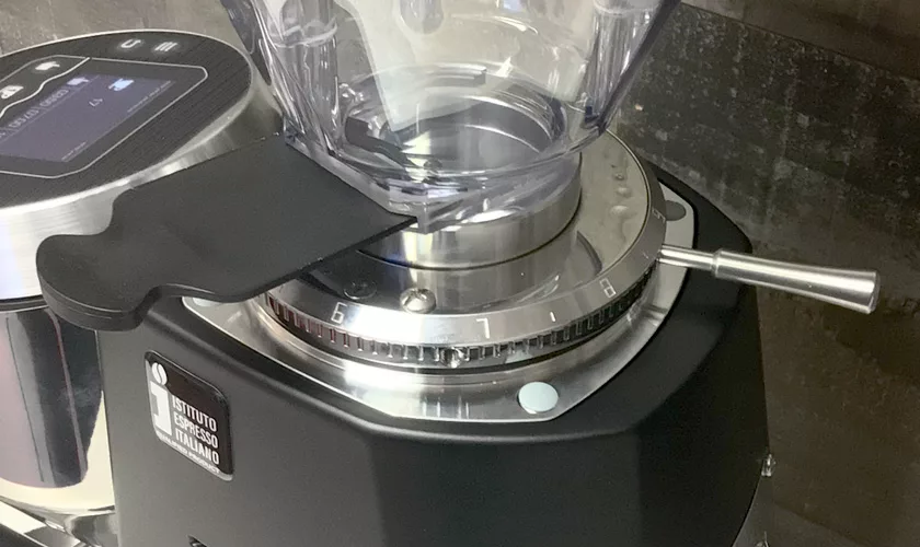 Close-up of a Mazzer Super Jolly V Pro Espresso Coffee Grinder featuring a transparent hopper, numbered metal adjustment dial, black plastic tool, and side metallic lever, set against a dark background. © 2025 1st-line.com